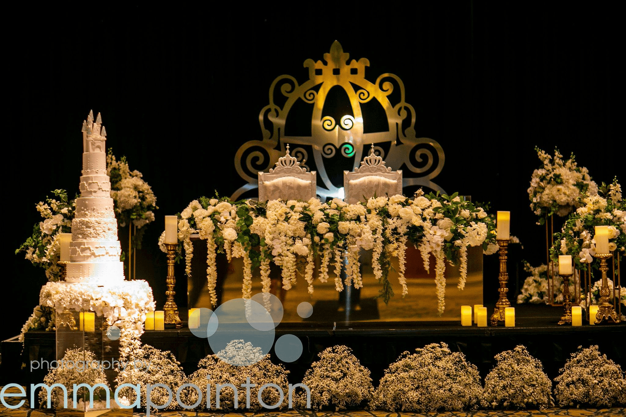 Reception stage backdrop with a gold carriage-crest design, a tall tiered wedding cake and candlelit floral arrangements