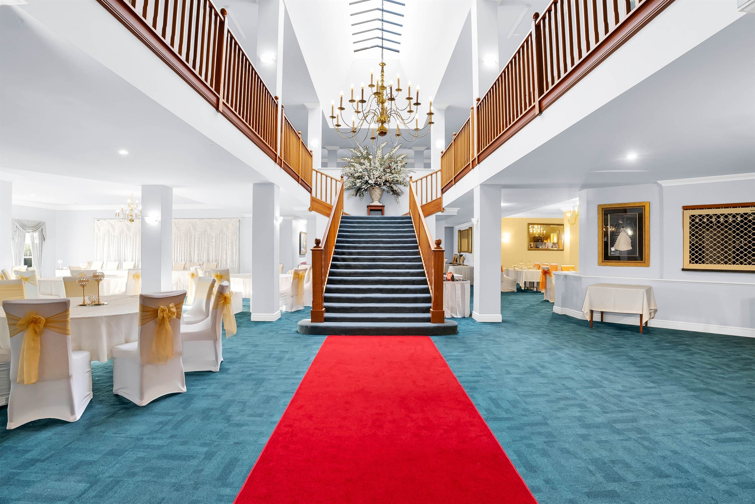 Grand foyer staircase and chandelier with a red carpet runner, flanked by round tables set with gold-sashed chair covers