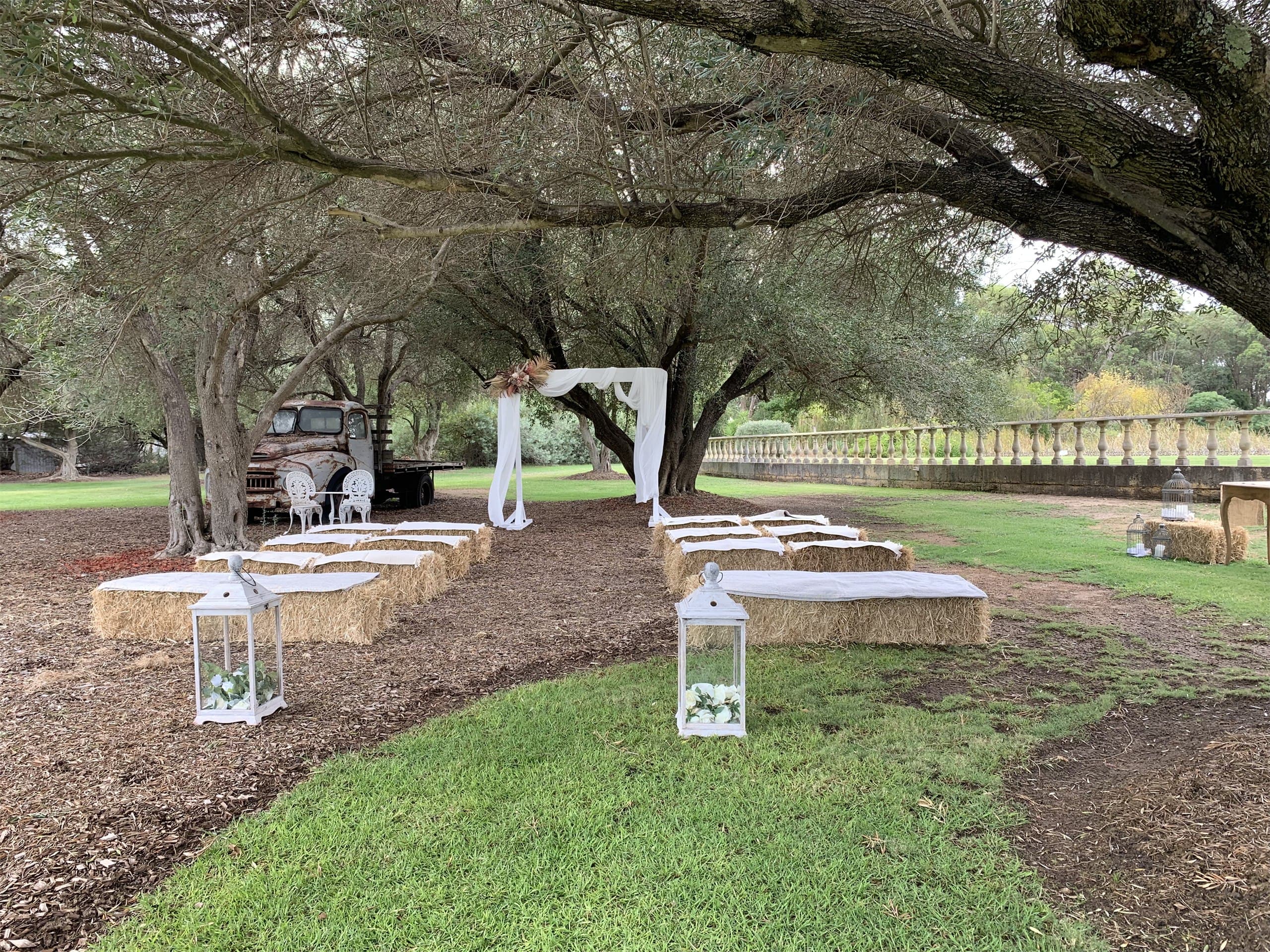 Rustic ceremony set-up under shade trees with a vintage flatbed truck, hay bale seating and a white drape arch
