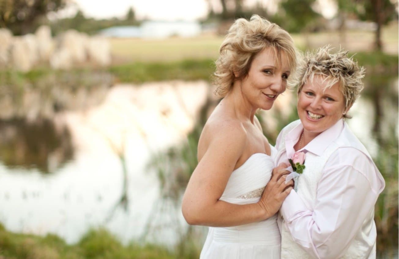 Two brides embracing beside the garden pond, one in a white strapless gown and one in a blush shirt with a boutonniere