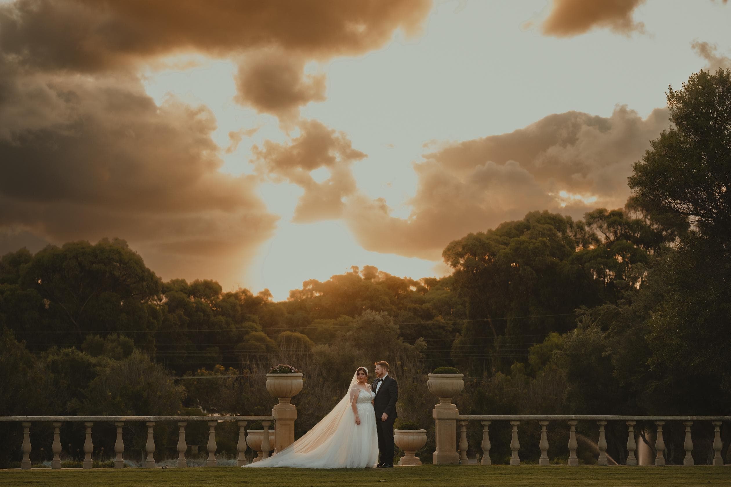 Bride and groom standing at the stone balustrade terrace under a dramatic orange-lit sunset sky