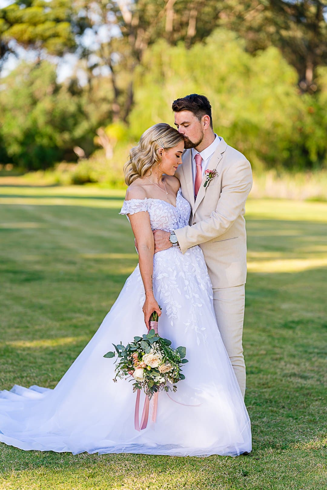 Groom kissing the bride's forehead on the lawn, bride holding a garden-style bouquet with trailing pink ribbon