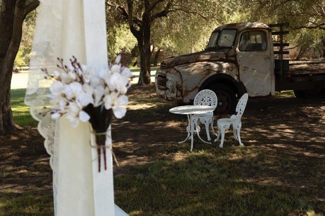 Dried floral bunch tied to a lace curtain, with a white wrought-iron bistro table and chairs beside the rustic truck ceremony set-up
