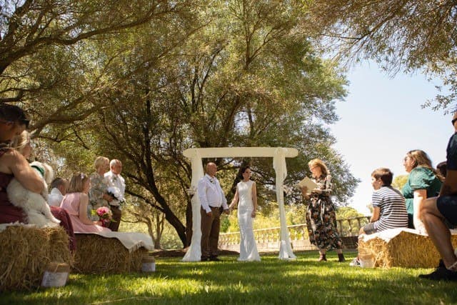 Celebrant leading a couple's vows under the trees, with guests seated on hay bale benches