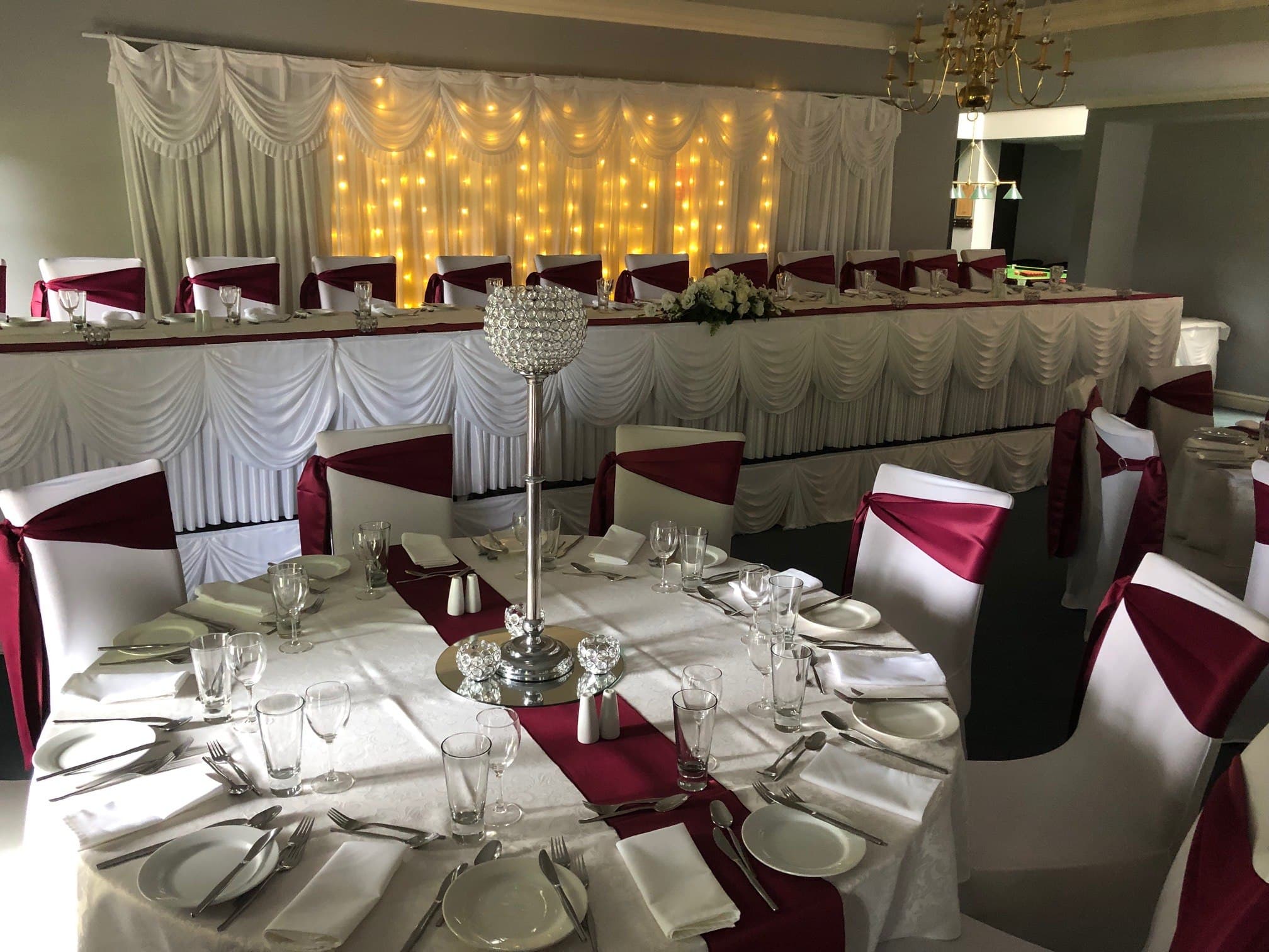 Head table with a fairy-light curtain backdrop, white swagged skirting and burgundy chair sashes
