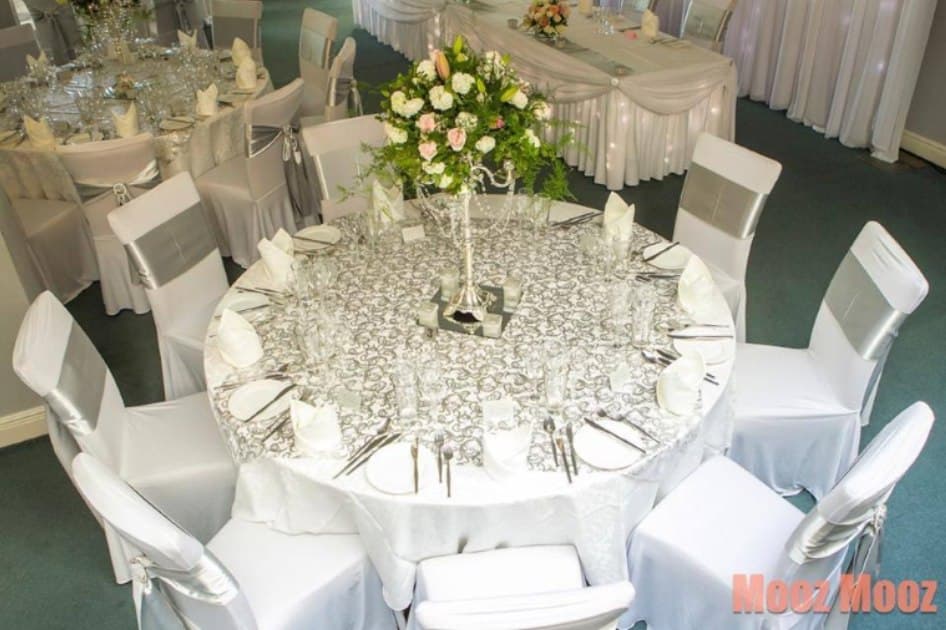 Round table with a silver patterned tablecloth, silver chair sashes and a crystal candelabra centrepiece